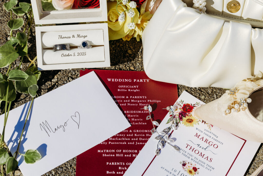 Close-up of wedding invitations and RSVP cards laid out with greenery and yellow flowers on white satin fabric.