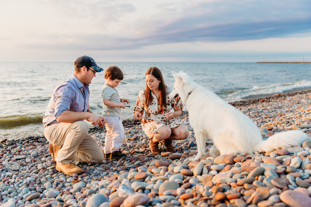 A family with a young child and a white dog sits on a pebbly beach by the sea during sunset, enjoying a calm, picturesque evening together.