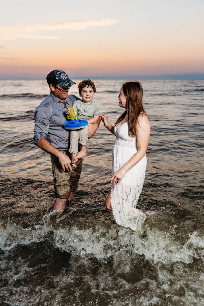 A family of three stands in the sea at sunset. Two adults smile at a child holding a toy boat. Waves and the beach surround them.