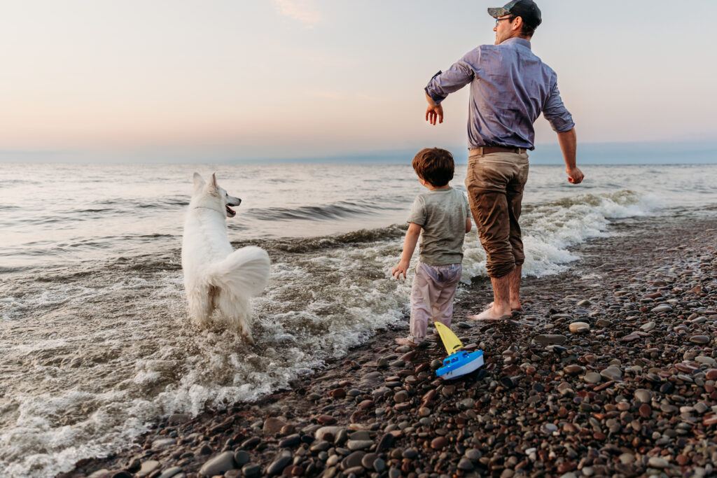 A person and a child walk along a pebble beach with a playful white dog; waves gently break, a toy boat rests on the shore, at dusk.