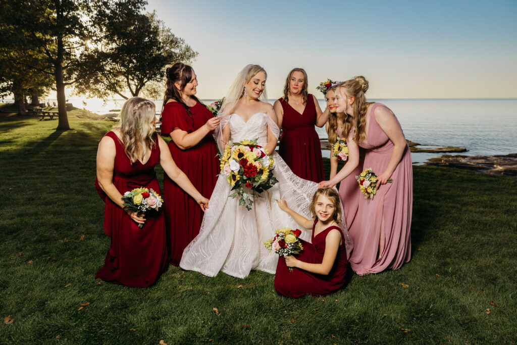 Bride in a white wedding gown stands with her bridesmaids in burgundy and blush dresses on a grassy lakeside.