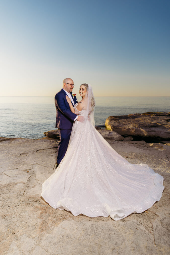 Bride and groom in wedding attire stand on a rocky shoreline by the sea, the bride's long train flowing behind them.