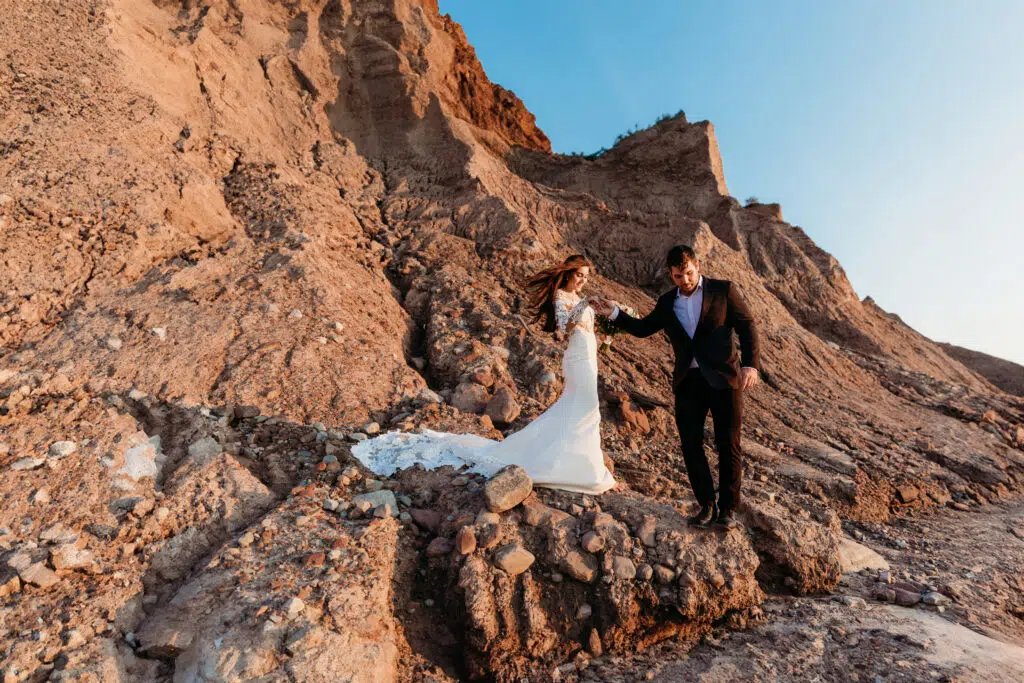 Two people in formal attire are walking hand in hand on a rocky slope at sunset, likely celebrating a wedding in a unique, rugged location.