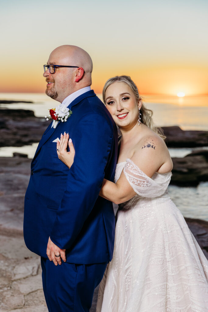 Bride in a white wedding dress hugs her groom in a blue suit at sunset on a rocky beach.