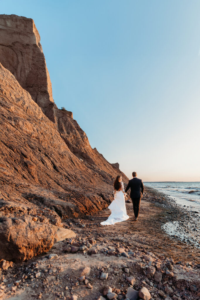 Two people, likely a couple, walk along a beach beside towering cliffs during golden hour, with one person wearing a white dress and the other in black.