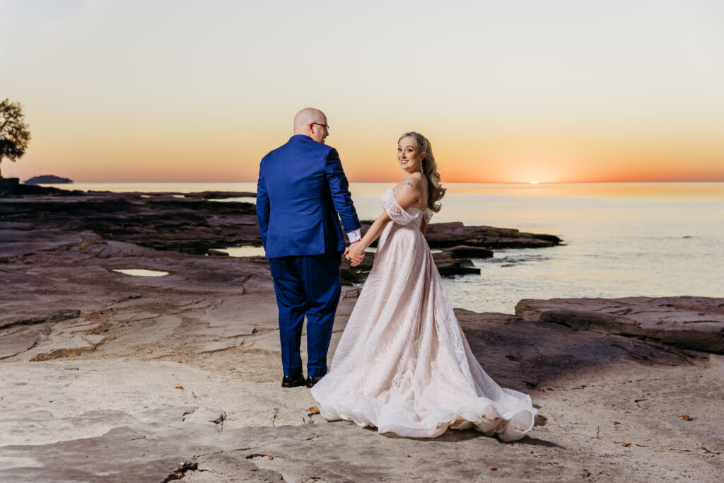 A bride in a white wedding gown and a groom in a blue suit stand on a rocky shoreline at sunset.