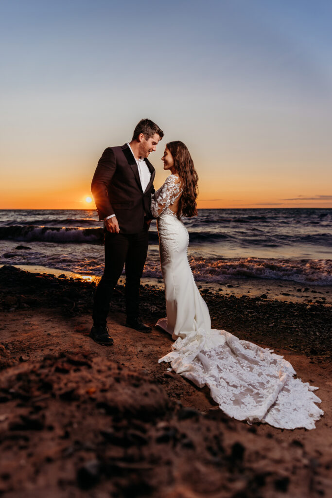 Two people stand on a beach at sunset, sharing a romantic moment. Person in a suit, person in a wedding dress with a long train. Waves and orange sky.