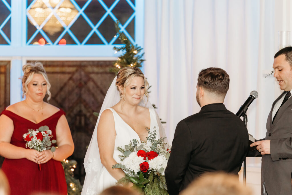 Three people at a wedding ceremony. One holds a bouquet, another reads from a microphone. Stained glass window in the background.