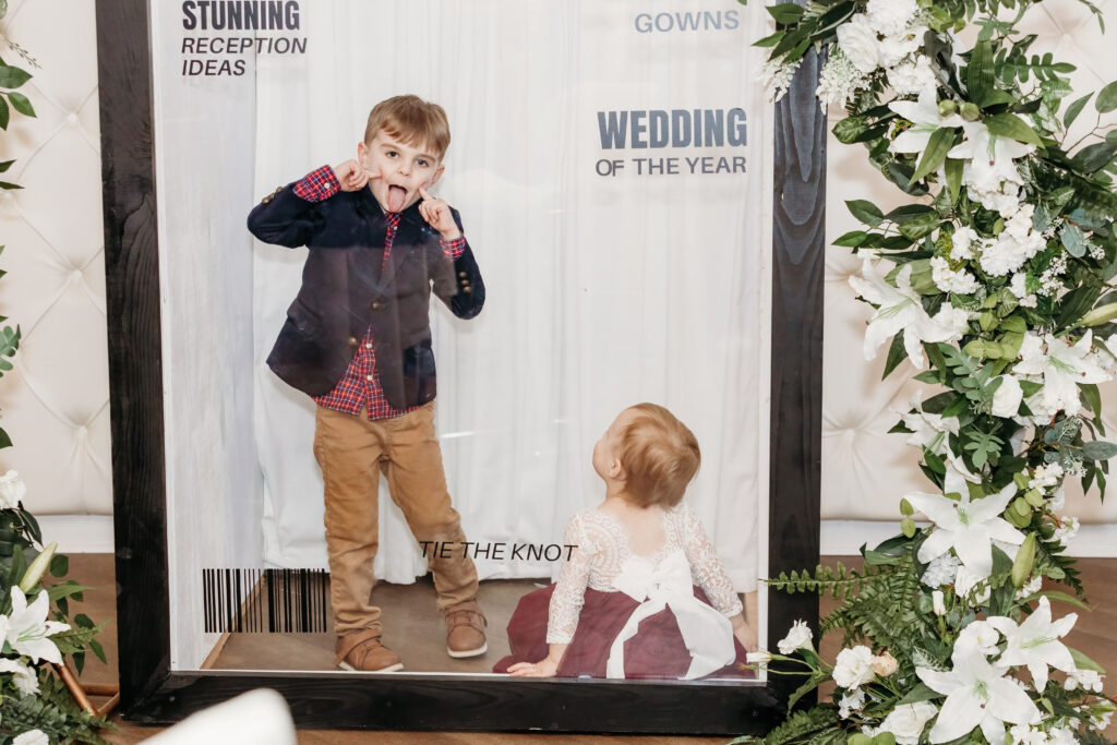 Two kids posing playfully inside a photo frame at a wedding event with floral decorations around the frame.