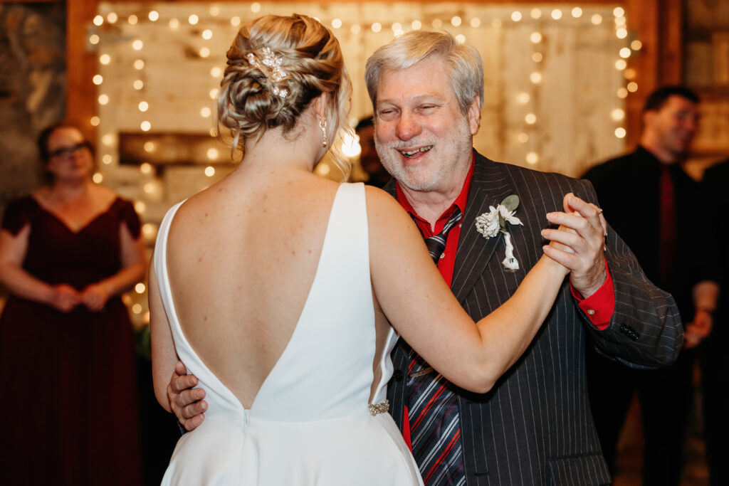 A person in a white dress dances joyfully with another person in a suit, amidst a festive setting with warm, twinkling lights.