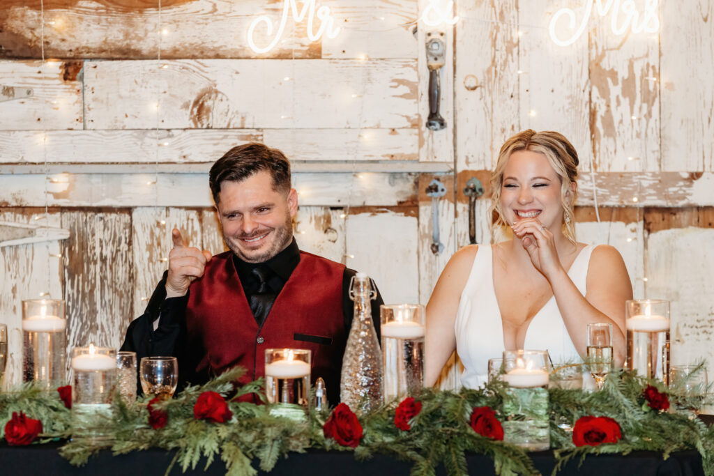Two people sit at a decorated table with candles and roses, smiling in a rustic setting with a