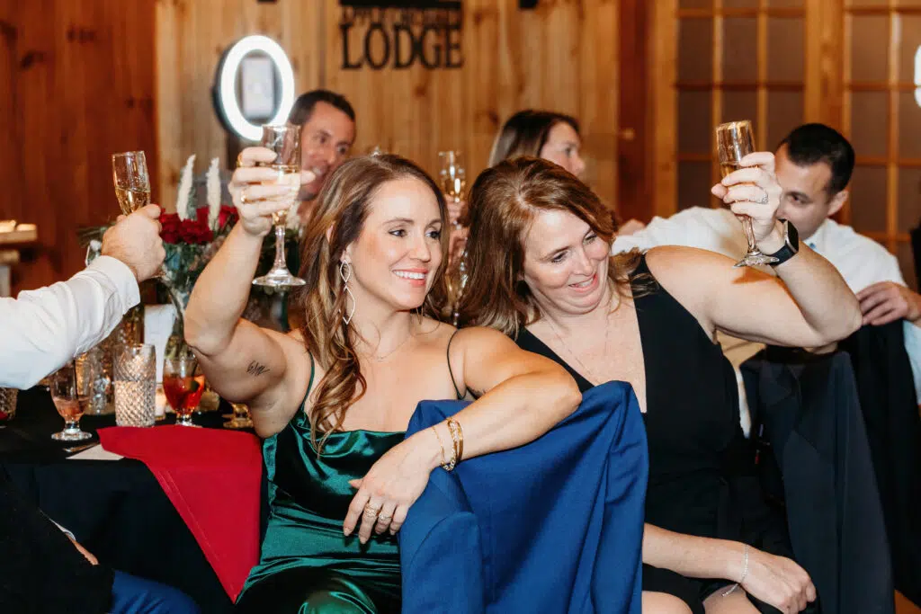 Two people in formal attire raise champagne glasses in a celebratory toast, surrounded by others at a warmly lit indoor gathering.
