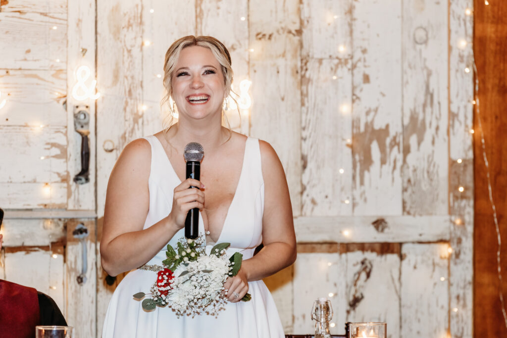 A person in a white dress holds a microphone, smiling and standing in front of rustic wooden doors with fairy lights.