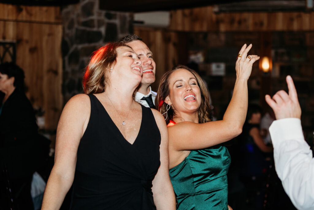 Three people are joyfully dancing together indoors, with wooden and stone wall decor in the background, creating a warm and lively atmosphere.