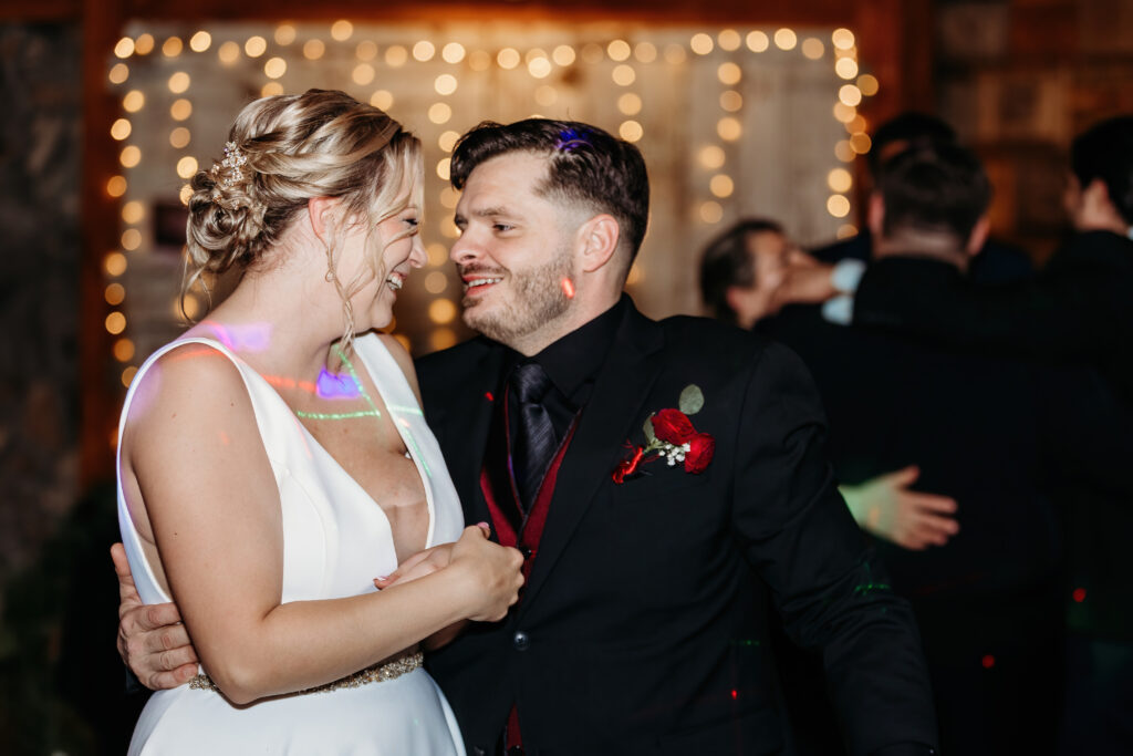 Two people joyfully dance at a festive event, surrounded by twinkling lights. Others are seen in the background, celebrating together.