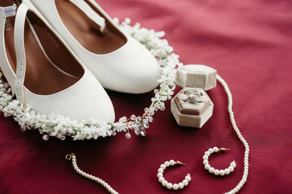 White shoes, a floral crown, a ring in a box, and pearl jewelry, are elegantly displayed on a crimson background.