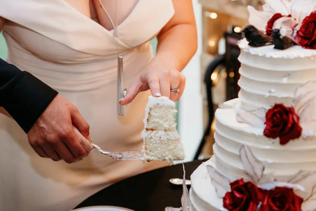 Two people are cutting a tiered white wedding cake decorated with red roses. One person is holding the slice, the other assisting.