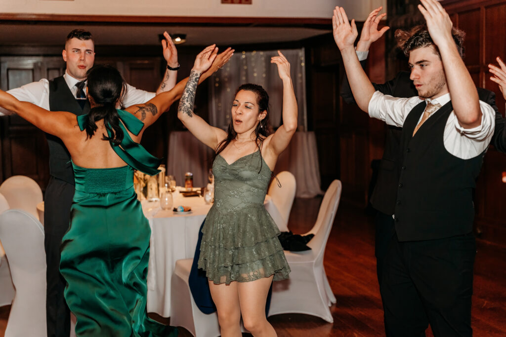Four people dance with raised arms at a formal indoor event, including two women in green dresses and two men in suits.