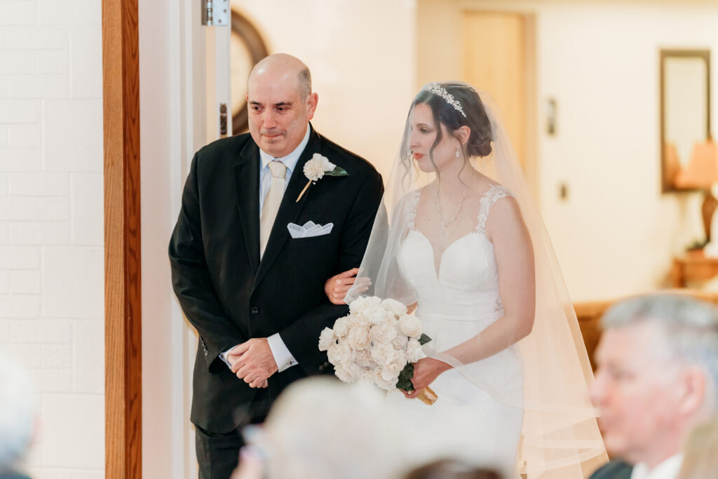 A bride in a white wedding gown and veil, holding a bouquet, walks down a hallway escorted by a man in a dark suit.