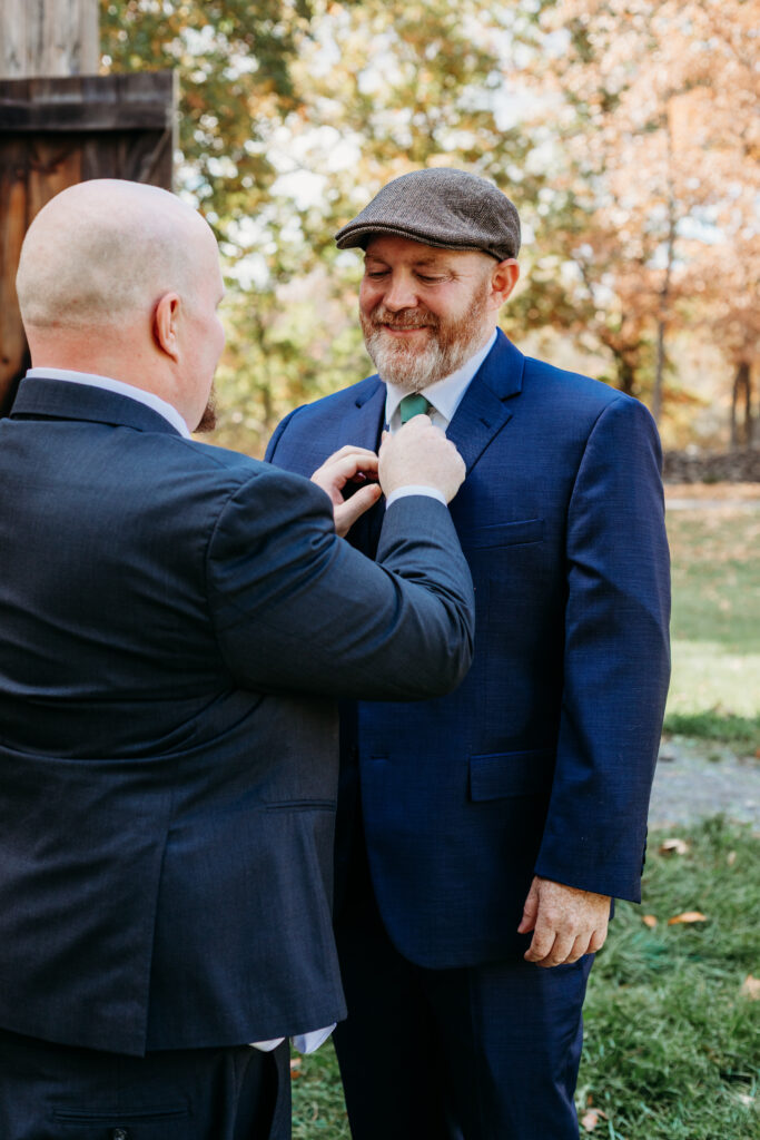 Two men in suits outdoors in autumn, with one pinning a boutonniere to the other’s lapel.