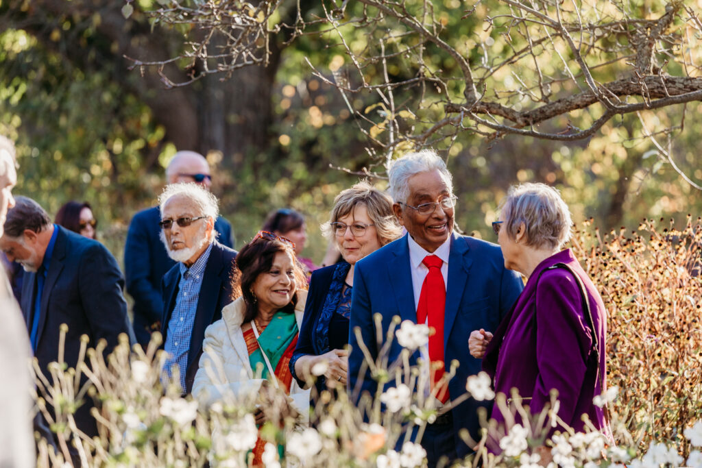A diverse group of well-dressed adults chat and smile outdoors among tall grasses and trees.