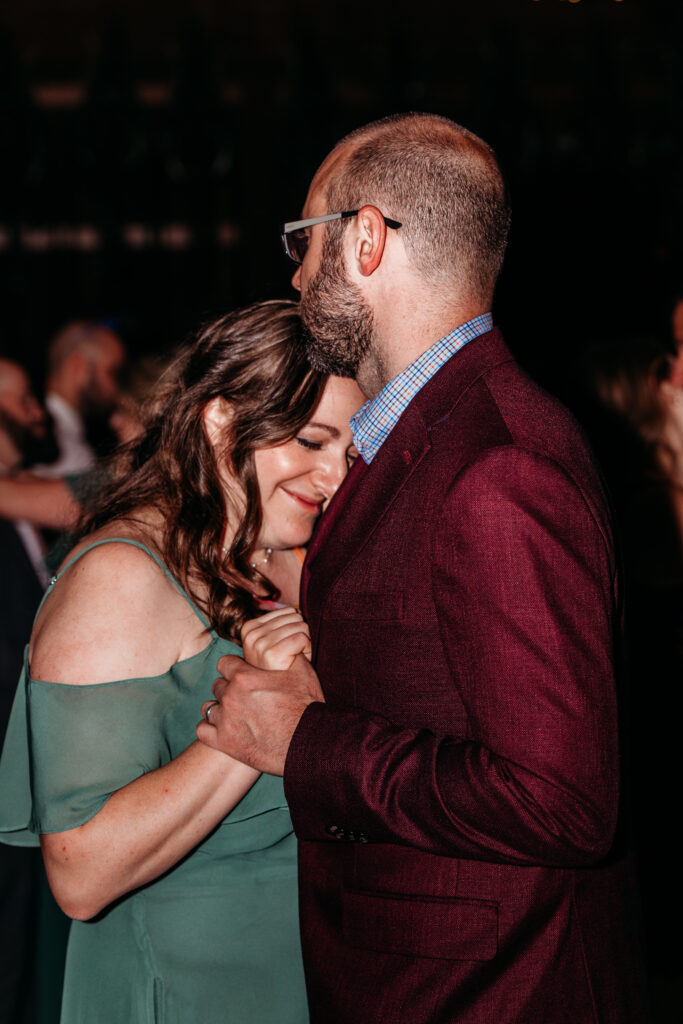A couple shares a tender hug at a dimly lit event, the woman wearing an off-the-shoulder green dress and the man in a burgundy suit.