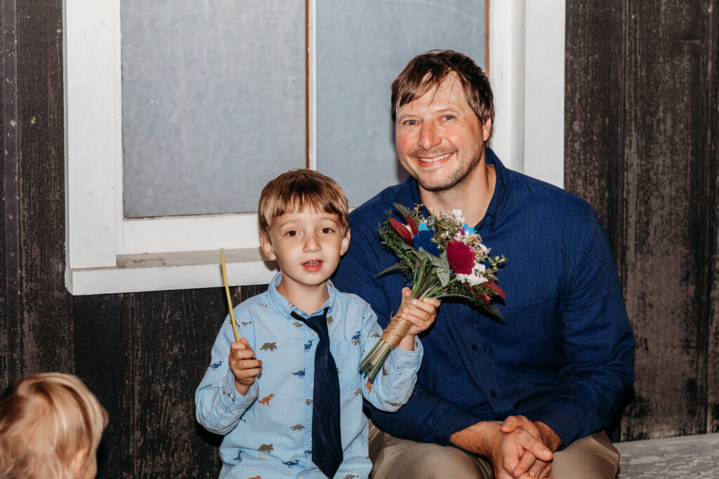 A man and a young boy sit together indoors, the boy holding a small candle and the man holding a bouquet of flowers.