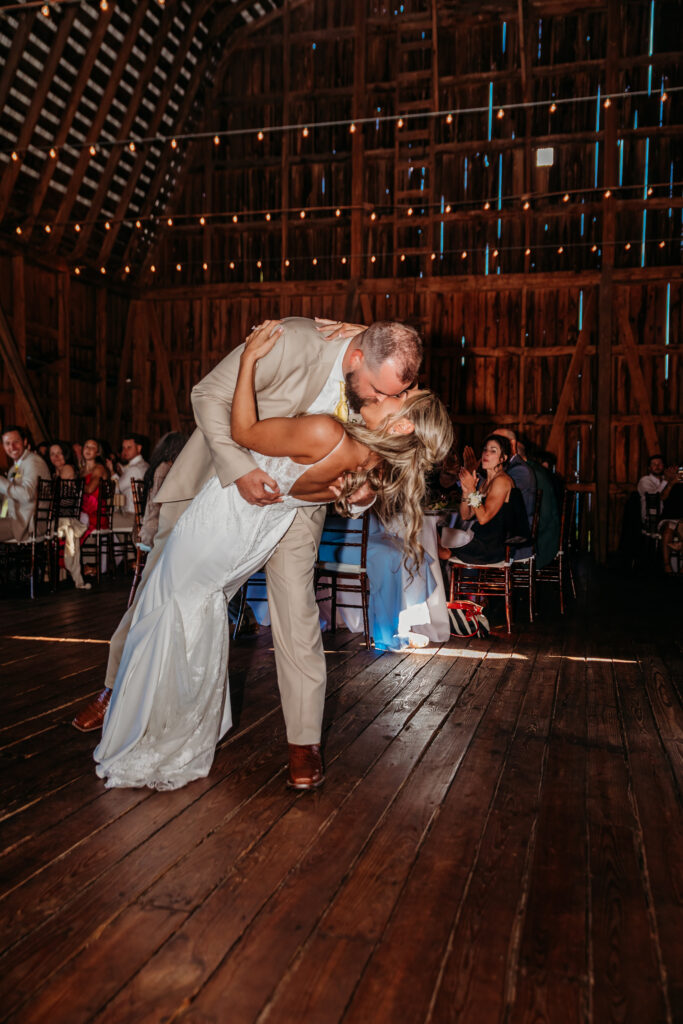 Bride and groom share a kiss while dancing on a wooden floor at a rustic barn wedding.