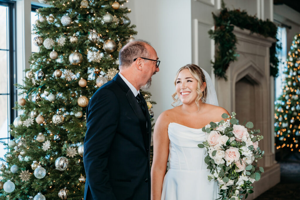 Bride in a white wedding dress holding a bouquet smiles at a groom in a dark suit beside a decorated Christmas tree.