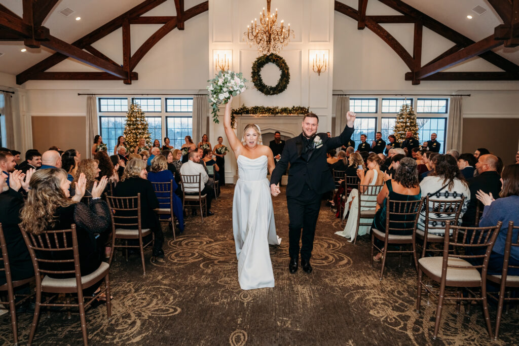 Bride and groom walk down the aisle with arms raised in celebration, surrounded by seated guests in a rustic venue with wooden beams and chandeliers.