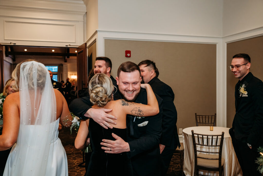 Wedding guests share a group hug in a hallway, with the bride in a white dress and veil standing nearby.