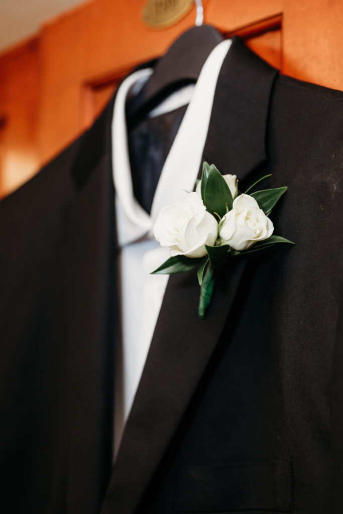 A black suit with a white rose boutonniere is neatly hung on a door, ready for a formal occasion.