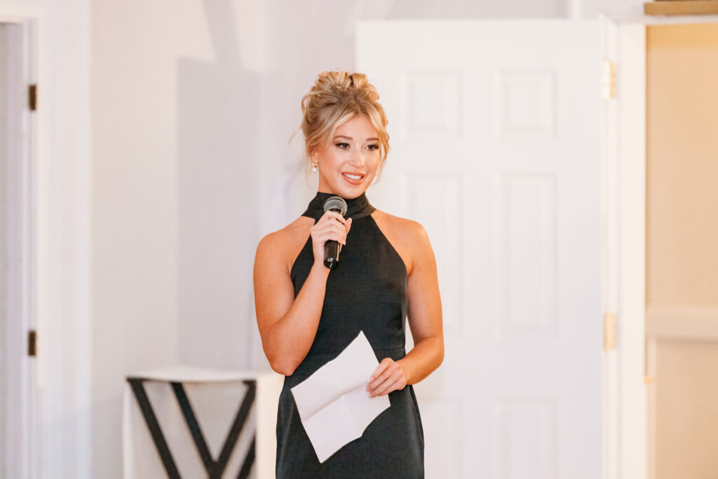 A person in a black dress holds a microphone and paper, standing indoors against a white background with doors visible.