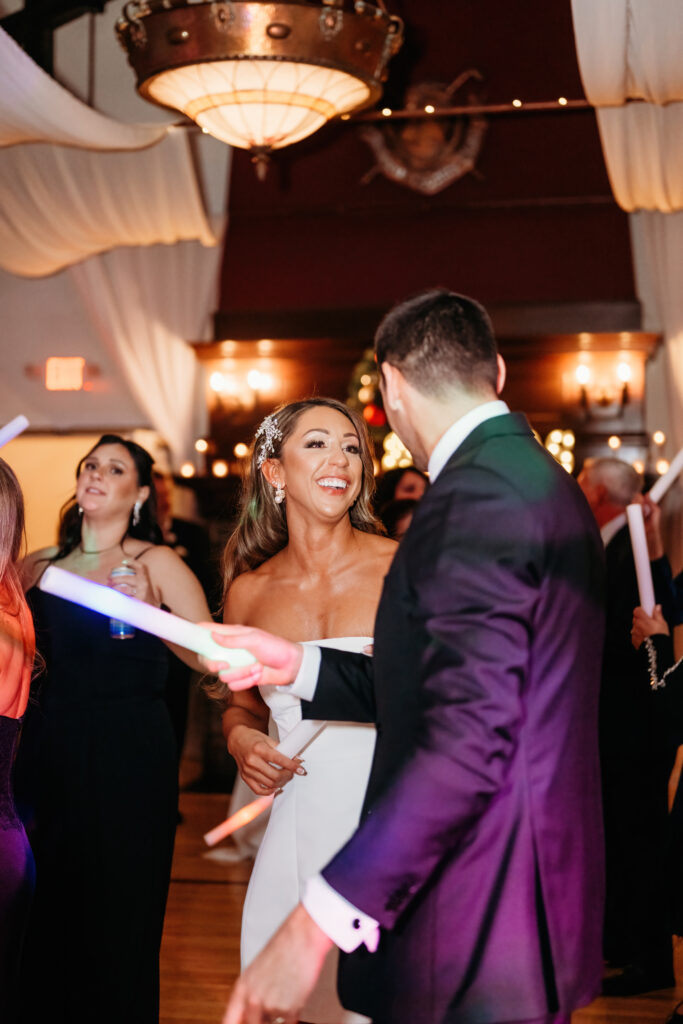 People dancing at an indoor event with elegant lighting and decor. Guests hold glow sticks, creating a festive atmosphere.
