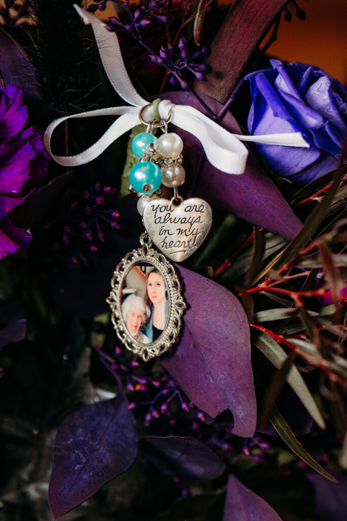 An ornate cameo-style oval locket containing a portrait, adorned with turquoise beads and a heart charm, suspended from a white ribbon among dark purple foliage.