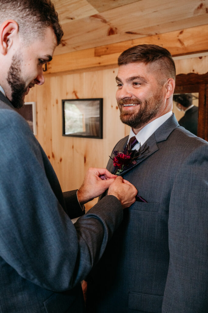 Two men in suits in a wood-paneled room; one pins a boutonniere on the other's lapel.