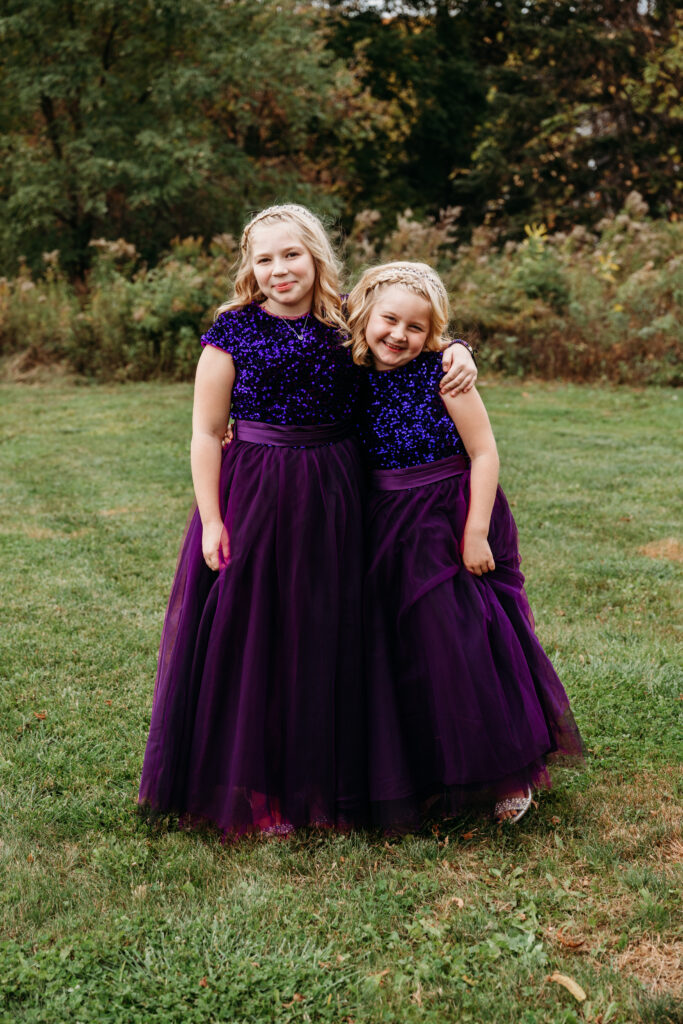 Two girls in matching purple dresses stand together on a grassy field with trees in the background.