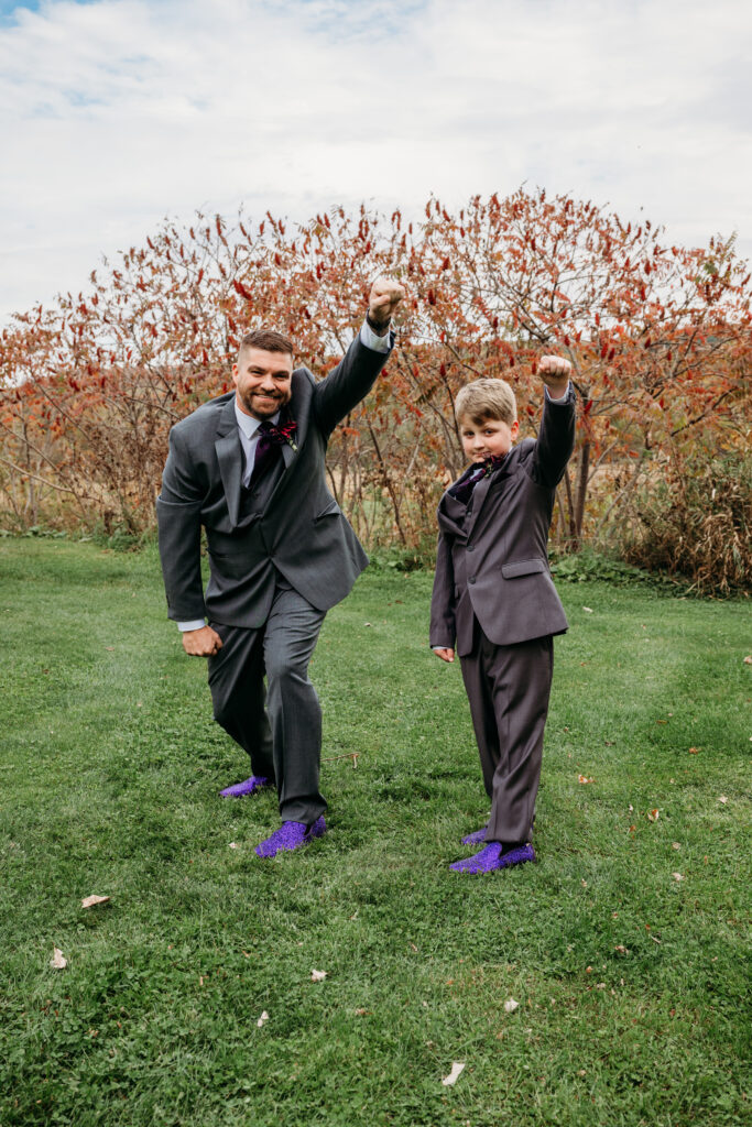 Two men in suits with purple shoes pose playfully in a grassy park with autumn trees in the background.