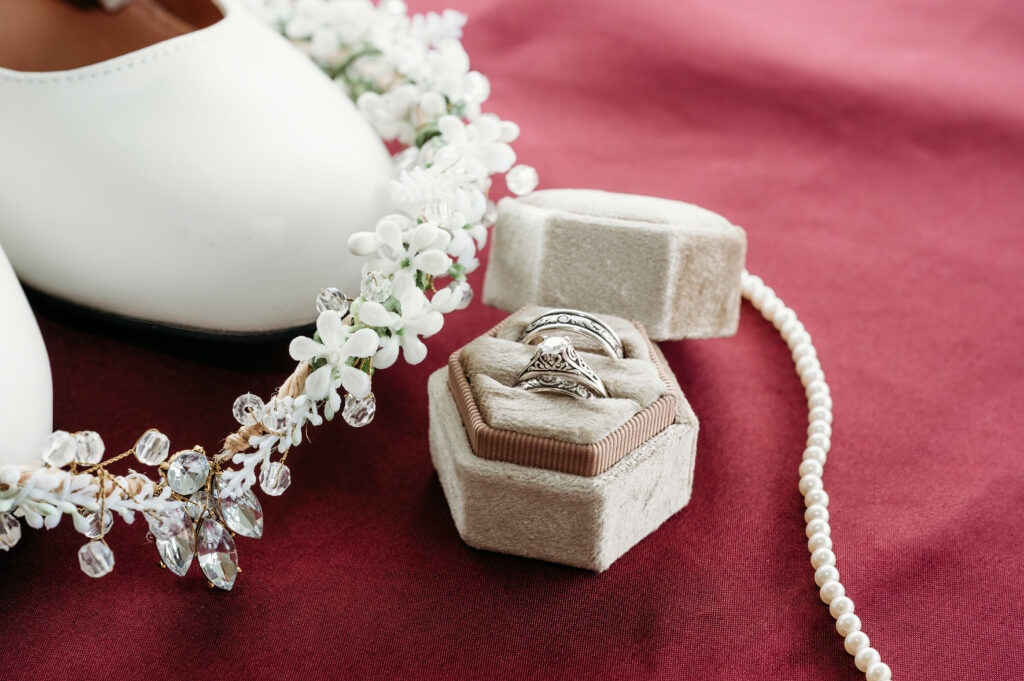 White shoes, pearl necklace, floral headpiece, and intricate silver rings in a velvet box are arranged on a burgundy fabric background.