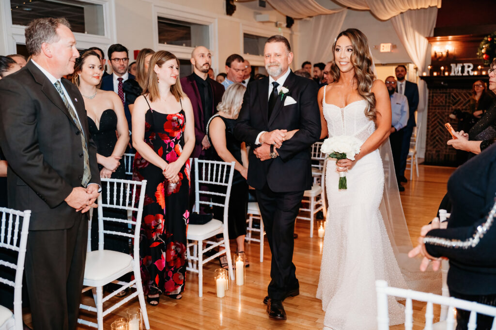 A wedding ceremony with a person walking another down the aisle, surrounded by seated guests in formal attire, on a wooden floor.
