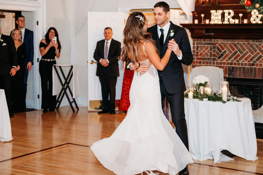 A couple dances at a wedding reception, surrounded by guests. Decor includes candles and greenery on tables with "MR." and "&" signs.