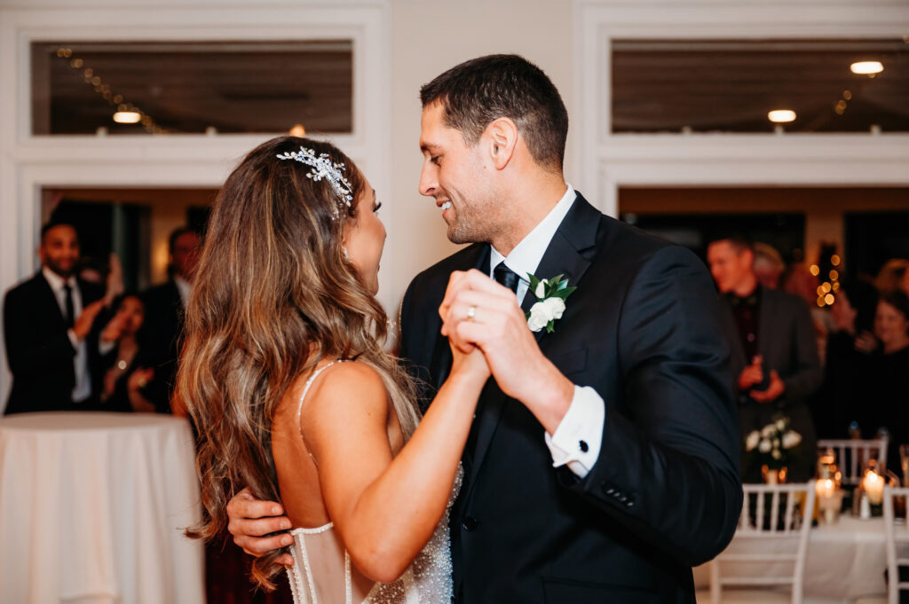 Two people dance closely at an indoor event. The person on the left has a decorative hairpiece, and guests are visible in the warmly lit background.