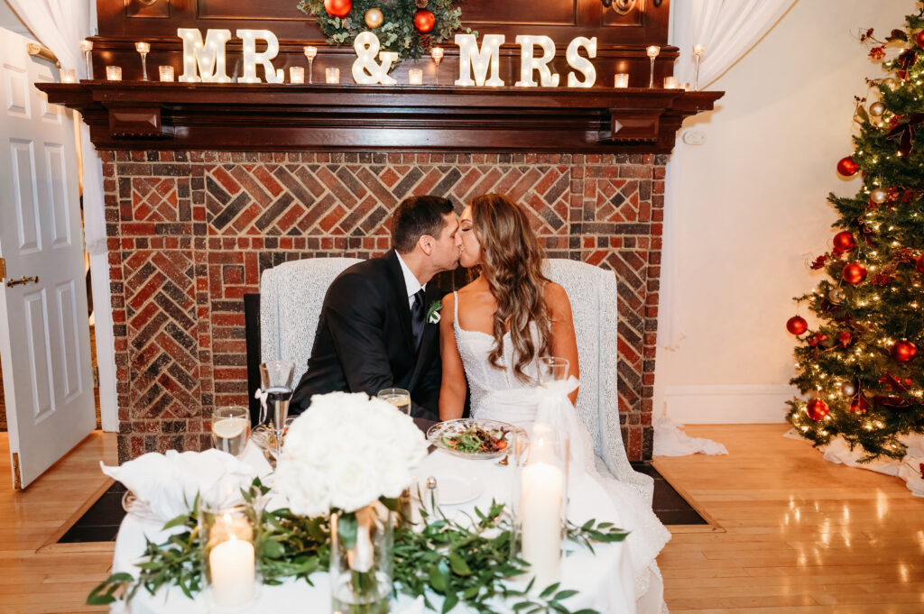 A couple kisses in front of a fireplace with "Mr. & Mrs." sign, surrounded by candles, floral centerpiece, and decorated Christmas tree.