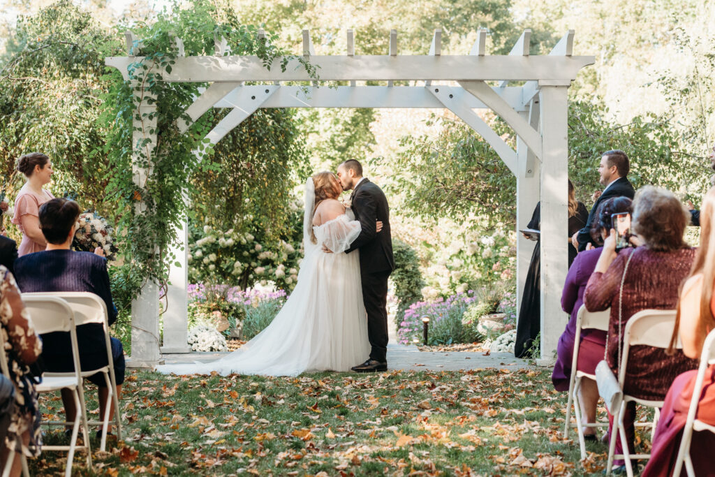 Bride and groom kiss under a white wooden arch at an outdoor garden wedding, with seated guests and fallen leaves.