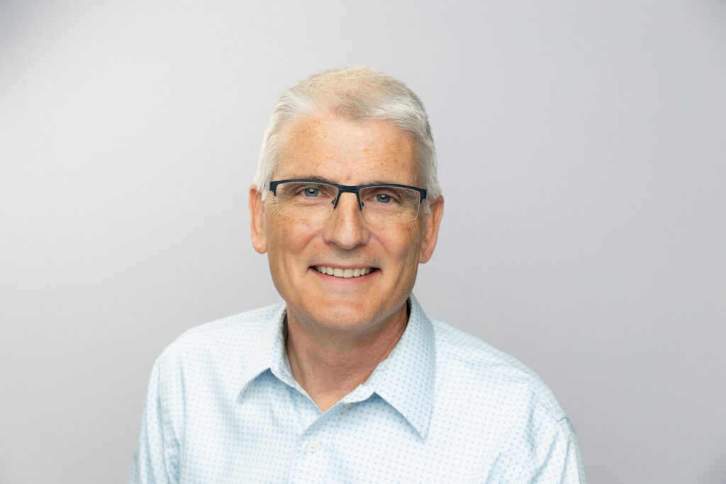 A portrait of a smiling person with white hair, wearing glasses, a light blue patterned shirt, against a plain light gray background.