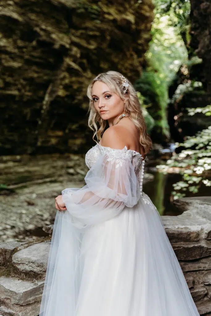 A person in a white bridal gown poses outdoors, looking over their shoulder, with a rocky backdrop and lush greenery surrounding them.