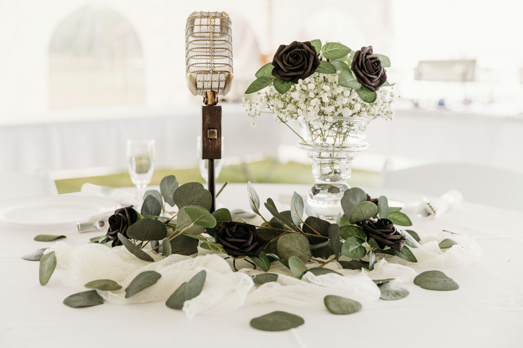 A vintage microphone stands atop a table with dark roses, green foliage, delicate white baby's breath in a vase, and clear glasses in the background.