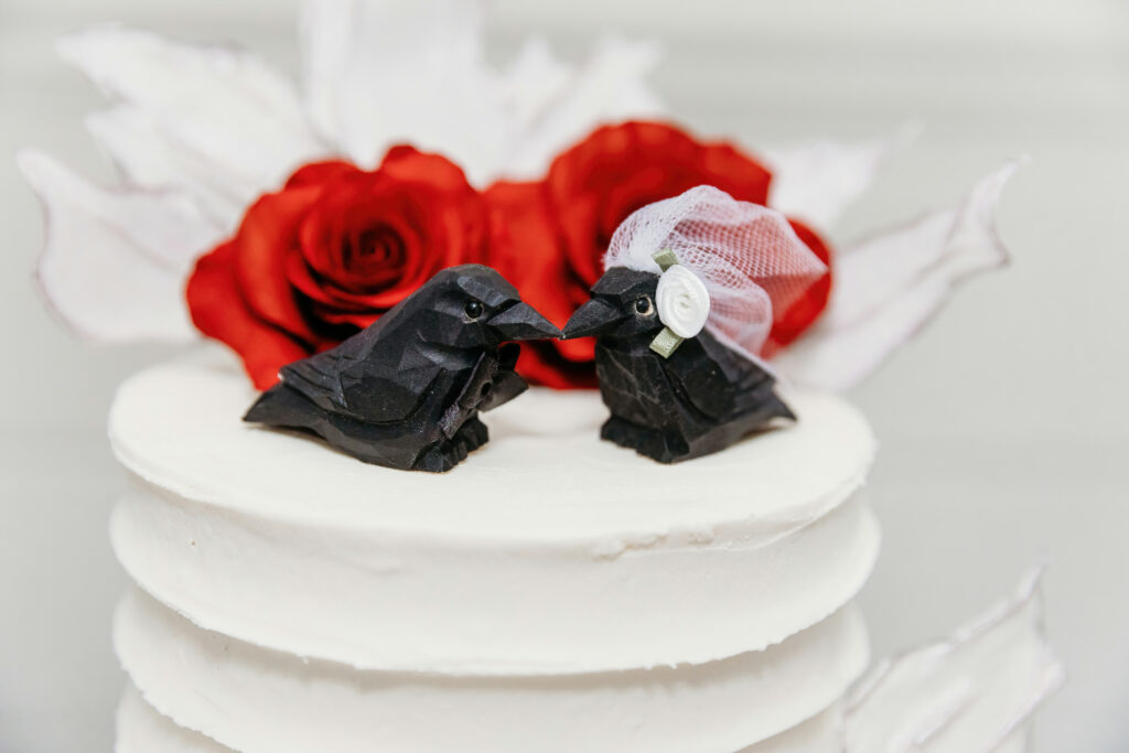 This image features two origami birds dressed as a bride and groom on top of a white cake, flanked by vibrant red roses and white petals.