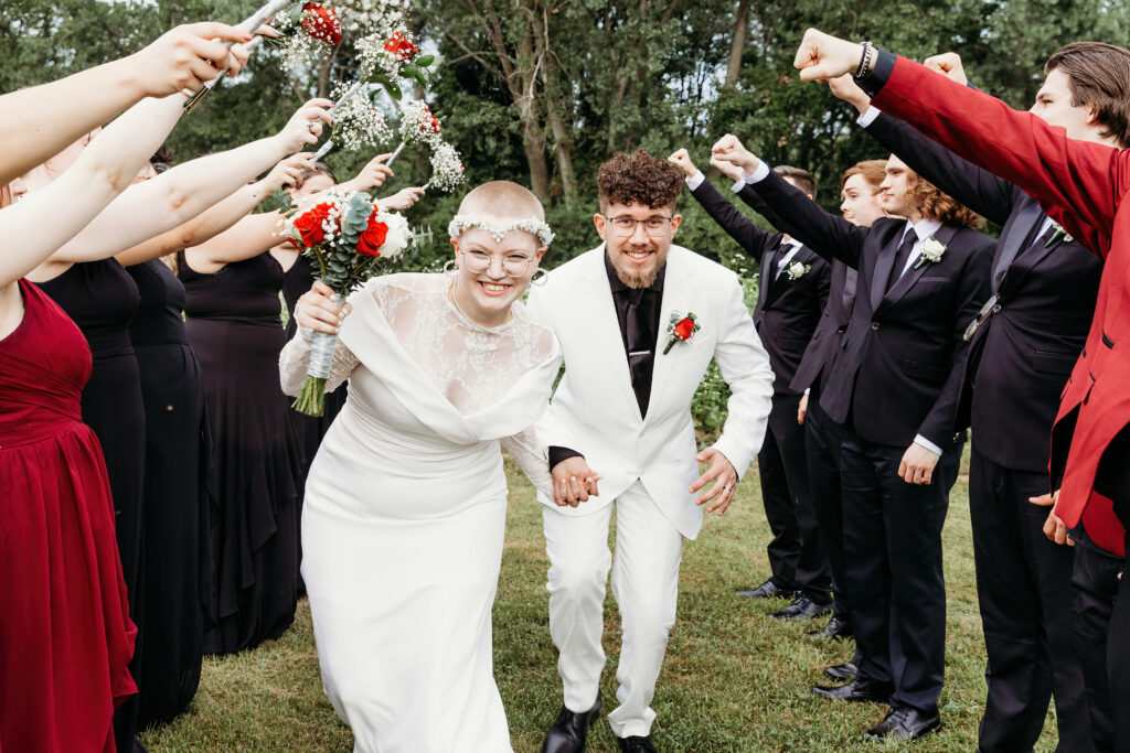Two joyful persons in wedding attire walk hand in hand under an arch of flowers held by well-dressed guests against an outdoor backdrop.
