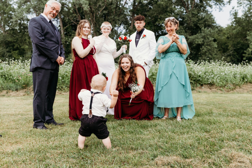 A toddler in a suspender outfit stands facing adults dressed in formal attire. They are smiling during a sunny outdoor event, likely a wedding.